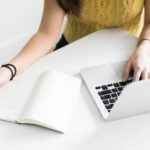 Hands on a laptop with a book nearby, wearing a yellow sweater, symbolizing HVAC study and productivity.
