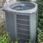 Gray air conditioning unit with a circular fan near greenery and a stone wall from AirLux Heating & Cooling.