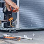 Hand holding a flame near refrigeration unit during HVAC repair, showcasing tools like wrenches and copper tubing.